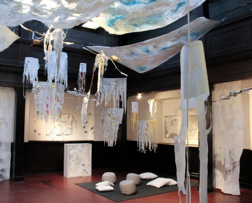 Lichte Kunstinstallation in einem holzgetäfelten Raum: Weiße Vliesbahnen und quallenartige Papierobjekte hängen über einer gemütlichen Sitzecke mit grauen Kissen.